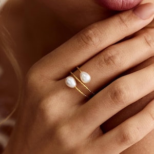 Könnte beinhalten: Ein goldener Ring mit zwei weißen, tropfenförmigen Perlen. Der Ring hat ein zartes, gedrehtes Banddesign. Die Hand ist im Fokus, wobei der Ring prominent auf der Haut dargestellt wird.