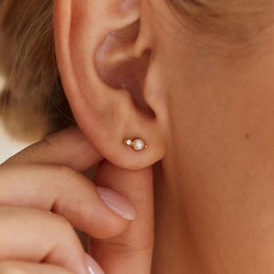 May include: A small gold stud earring featuring a tiny white pearl and a single clear gemstone set in a horizontal bar design. The earring is positioned on the earlobe, showcasing a delicate metallic finish and a simple, refined structure.