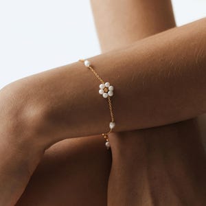 Peut inclure: Un bracelet délicat en chaîne dorée orné d'un motif floral composé de six perles blanches avec une perle centrale dorée, agrémenté de perles blanches isolées espacées le long de la fine chaîne à maillons métalliques.