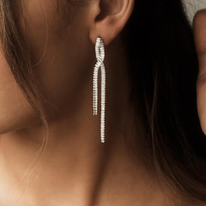 May include: Elegant silver earrings with a linear design. The earrings feature a twisted top section and a long, dangling, linear bottom section. The earrings are adorned with small, sparkling stones, creating a glamorous look.