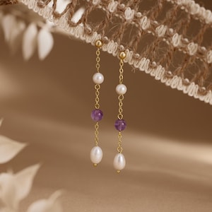 May include: A pair of gold-toned drop earrings featuring a delicate chain design. Each earring is accented with a small white round pearl, a purple spherical gemstone, and a larger white teardrop-shaped pearl at the base of the chain.
