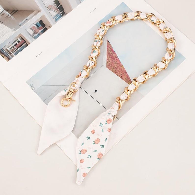 May include: A gold chain strap with white fabric woven through the links. The fabric features a floral pattern in pink and green. The strap has gold-coloured clasps on each end.