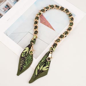May include: A gold-coloured chain bag strap with olive green fabric woven through the links. The strap ends in olive green fabric with a fern leaf pattern and gold-coloured clasps.