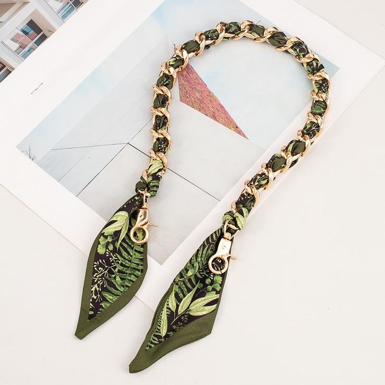 May include: A gold-coloured chain bag strap with olive green fabric woven through the links. The strap ends in olive green fabric with a fern leaf pattern and gold-coloured clasps.