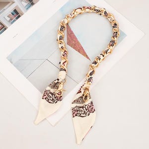 May include: A cream-coloured purse strap with a gold chain and patterned silk scarf accents. The scarf features a black and pink design. The strap has gold-coloured clasps for attachment.