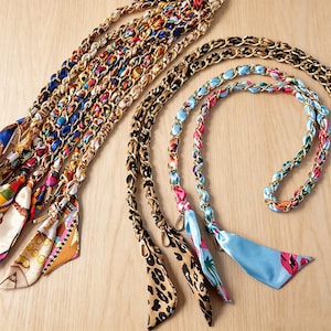May include: A collection of bag straps featuring gold-tone metal chains interwoven with colorful patterned fabric ribbons. The straps display various designs including leopard prints, tropical motifs, and geometric patterns, each finished with metal clasps.