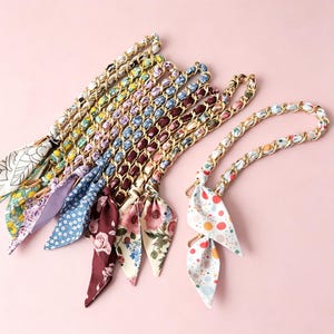 May include: A collection of bag straps featuring gold-tone metal chains interwoven with various patterned fabric ribbons. The ribbons display diverse designs including floral prints, polka dots, and abstract motifs, each ending in a pointed tail.