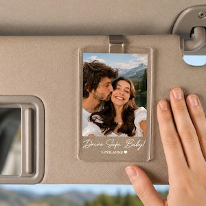 Puede incluir: Un clip rectangular de acrílico transparente para el parasol del coche con una fotografía en color de una pareja al aire libre con montañas de fondo. El texto "Drive Safe, Baby!" y "LOVE, ANNE" con un icono de corazón está impreso en blanco en la parte inferior.