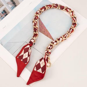 May include: A handbag strap with a gold chain and red fabric. The chain is interwoven with red fabric, and the ends have red diamond-patterned fabric. The strap has gold clasps for attachment.