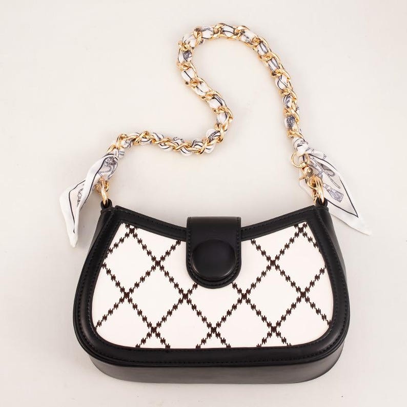 May include: A black and white handbag with a chain strap and a small scarf detail. The bag features a white front panel with a black diamond pattern, a black flap closure, and a gold chain strap.