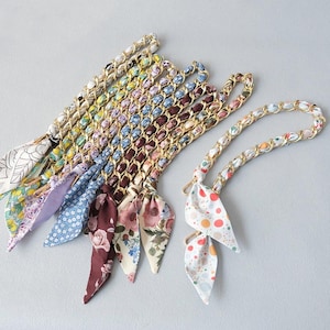 May include: A selection of patterned silk scarf handbag straps with gold-toned chain links. The scarves display floral and geometric patterns in various colours, including blue, pink, yellow, and white. The straps are designed for handbags.
