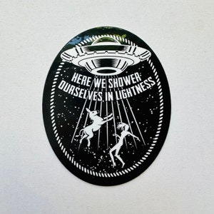 Può includere: Adesivo bianco e nero con un UFO, un asino e una persona che viene sollevata da raggi di luce. Il testo dice "Here we shower ourselves in lightness."