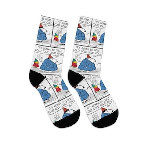 May include: Pair of white socks featuring a comic strip with Snoopy and Woodstock in winter attire. The text reads "BE COLD, COLD, COLD, COLD" and "IT'S GONNA BE COLD, COLD, COLD, COLD." Black heels and toes complete the design.