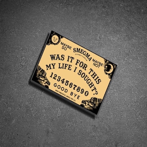 May include: A black and gold Ouija board with the text "SMEGMA DOGMATAGRAA" and "WAS IT FOR THIS MY LIFE I SOUGHT?"  The board also has the numbers 1234567890 and the words "GOOD BYE".