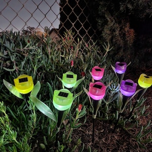 Puede incluir: Un conjunto de siete luces solares de flores de colores. Las luces tienen forma de tulipanes y están hechas de plástico. Están iluminadas con LED de diferentes colores. Las luces están dispuestas en un entorno de jardín.