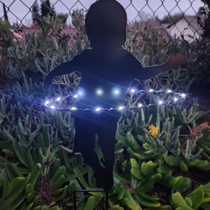 May include: A black metal silhouette of a child jumping rope with white LED lights around the rope. The silhouette is placed in a garden setting with green plants.