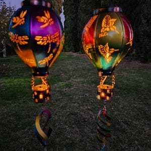 Puede incluir: Dos faroles solares coloridos con forma de globo aerostático con recortes de libélulas y colibríes. Los faroles tienen un degradado de colores, incluyendo morado, verde y naranja. Cada farol tiene una espiral decorativa colgante.