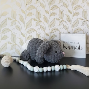 May include: A handmade gray crochet bunny with a white fluffy tail and pink nose. A wooden sign with the text "Happiness is homemade" is next to the bunny. A decorative bead garland with white, gray, and green beads is in the foreground.