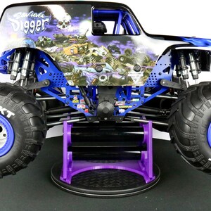 May include: A detailed image of a monster truck toy, featuring a black and blue body with the words "Grave Digger" and a skull graphic. The truck has large black tires with blue rims and is supported by a purple stand.