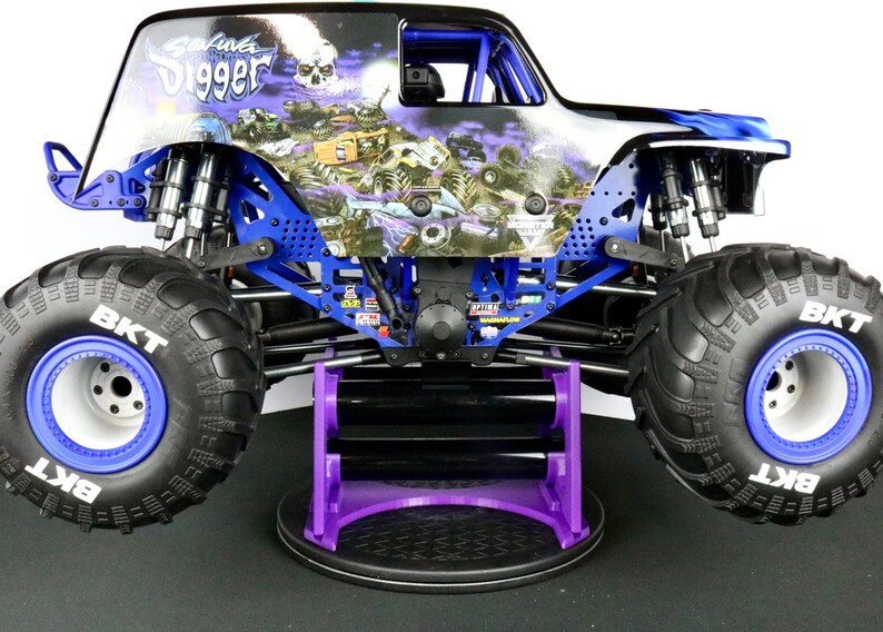 May include: A detailed image of a monster truck toy, featuring a black and blue body with the words "Grave Digger" and a skull graphic. The truck has large black tires with blue rims and is supported by a purple stand.