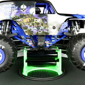 May include: A blue and white Grave Digger monster truck toy with large black tires and blue rims. The truck has a skull graphic on the side and is displayed on a green and black stand.