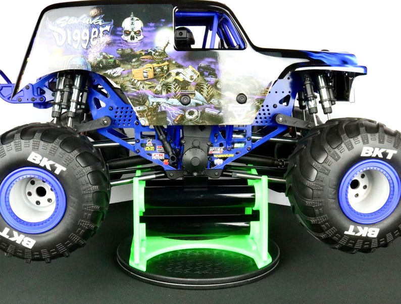 May include: A blue and white Grave Digger monster truck toy with large black tires and blue rims. The truck has a skull graphic on the side and is displayed on a green and black stand.