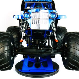 May include: A blue and black monster truck toy with large black tires. The truck has a blue and silver front with two round headlights. The truck is sitting on a blue stand.