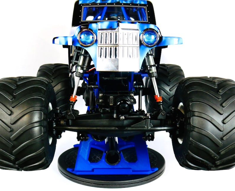 May include: A blue and black monster truck toy with large black tires. The truck has a blue and silver front with two round headlights. The truck is sitting on a blue stand.