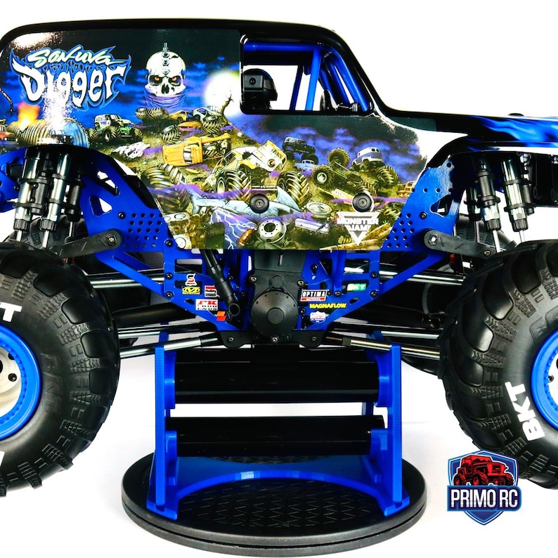 May include: A blue and black monster truck with large black tires and blue rims. The truck has a detailed graphic design with the words "Grave Digger" and a skull. The truck is on a blue stand.