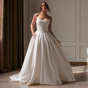 May include: A strapless ivory wedding dress featuring a fitted bodice with horizontal ruching and a sweetheart neckline. The gown has a full, floor-length skirt with a smooth satin finish and a subtle train, creating a classic and elegant silhouette.