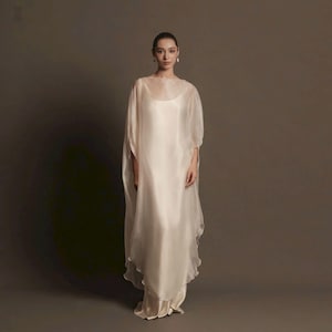 May include: A floor-length, cream-colored dress featuring a sleeveless, opaque inner layer and a sheer, flowing outer layer with wide, draped sleeves. The garment has a rounded neckline and a loose, relaxed silhouette that falls to the floor.