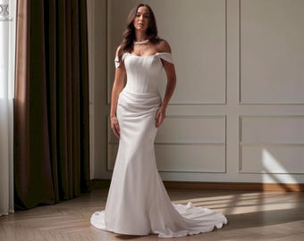 May include: A floor-length white gown featuring an off-the-shoulder neckline, a structured corset-style bodice with vertical seams, and a draped skirt that flows into a small train. The fabric has a smooth, matte finish throughout the garment.