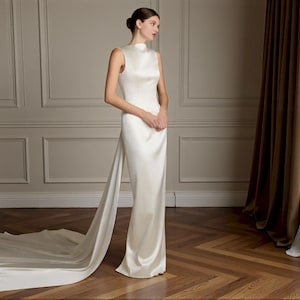 K&ouml;nnte beinhalten: Ein bodenlanges, elfenbeinfarbenes Brautkleid mit hohem Ausschnitt und Schleppe. Das Kleid ist aus einem glatten, satinartigen Stoff gefertigt. Das Model steht in einem Raum mit Parkettboden und neutralfarbenen W&auml;nden. Das Kleid hat ein schlichtes, elegantes Design.