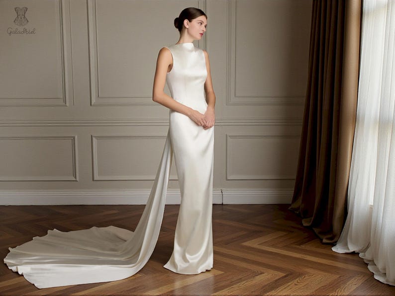 K&ouml;nnte beinhalten: Ein bodenlanges, elfenbeinfarbenes Brautkleid mit hohem Ausschnitt und Schleppe. Das Kleid ist aus einem glatten, satinartigen Stoff gefertigt. Das Model steht in einem Raum mit Parkettboden und neutralfarbenen W&auml;nden. Das Kleid hat ein schlichtes, elegantes Design.
