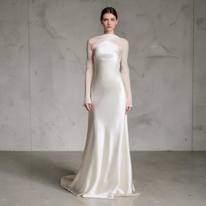 Puede incluir: Un vestido largo de satén color marfil con cuello alto y mangas largas y transparentes de color rosa pálido que se cruzan sobre el pecho. El vestido tiene un corpiño ajustado que fluye hacia una falda larga con una ligera cola.