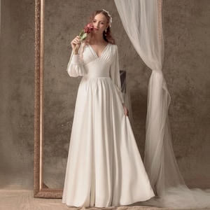 Puede incluir: Un vestido de novia de color marfil con escote en V y mangas largas. El vestido tiene una falda fluida. La imagen incluye un espejo decorativo y cortinas transparentes. La novia sostiene una flor. La estética general es elegante y romántica.