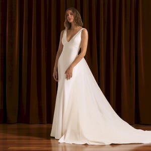 Puede incluir: Un vestido de novia blanco con escote en V y una larga cola. El vestido es largo hasta el suelo y est&aacute; hecho de una tela suave y elegante. La modelo est&aacute; de pie frente a una cortina marr&oacute;n oscuro.