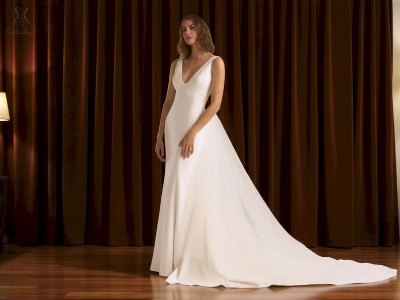 Puede incluir: Un vestido de novia blanco con escote en V y una larga cola. El vestido es largo hasta el suelo y est&aacute; hecho de una tela suave y elegante. La modelo est&aacute; de pie frente a una cortina marr&oacute;n oscuro.