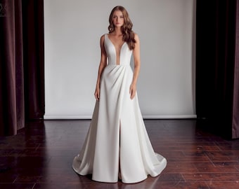 May include: A white sleeveless wedding gown with a plunging V-neckline and a high slit. The dress has a flowing A-line skirt and a long train. The model is standing in front of a white backdrop.