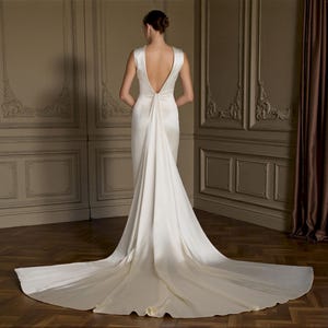 K&ouml;nnte beinhalten: R&uuml;ckansicht eines wei&szlig;en Brautkleides mit langer Schleppe. Das Kleid hat einen tiefen R&uuml;ckenausschnitt und ein schlichtes, elegantes Design. Die Braut steht in einem Raum mit Holzb&ouml;den und dekorativen Wandpaneelen.