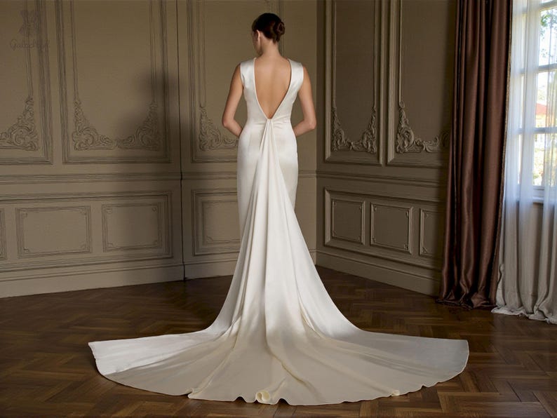 K&ouml;nnte beinhalten: R&uuml;ckansicht eines wei&szlig;en Brautkleides mit langer Schleppe. Das Kleid hat einen tiefen R&uuml;ckenausschnitt und ein schlichtes, elegantes Design. Die Braut steht in einem Raum mit Holzb&ouml;den und dekorativen Wandpaneelen.