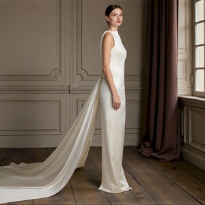 K&ouml;nnte beinhalten: Ein &auml;rmelloses, bodenlanges elfenbeinfarbenes Kleid mit langer Schleppe. Das Kleid hat einen hohen Ausschnitt und eine figurbetonte Silhouette. Die Schleppe erstreckt sich vom R&uuml;cken des Kleides und erzeugt einen dramatischen Effekt.