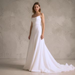 Könnte beinhalten: Ein weißes Brautkleid mit einer fließenden Schleppe. Das Kleid hat ein schlichtes, elegantes Design mit Spaghettiträgern und einem figurbetonten Oberteil. Das Model steht vor einer neutral getönten Wand.