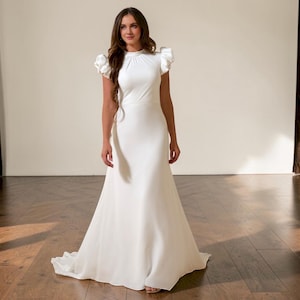 Könnte beinhalten: Ein weißes Brautkleid mit hohem Ausschnitt und kurzen, gerüschten Ärmeln. Das Kleid hat ein figurbetontes Oberteil und einen fließenden Rock, der sich ab der Taille ausstellt. Das bodenlange Kleid ist aus einem glatten, eleganten Stoff gefertigt.