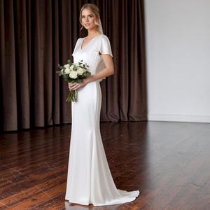 May include: A white wedding dress with a V-neckline and short sleeves. The dress has a fitted bodice and a flared skirt. The person is holding a bouquet of white flowers and wearing a veil. The background features brown curtains and a wooden floor.