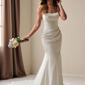 K&ouml;nnte beinhalten: Ein wei&szlig;es, bodenlanges Hochzeitskleid mit einer Meerjungfrau-Silhouette. Das Kleid hat einen quadratischen Ausschnitt, Spaghettitr&auml;ger und ein drapiertes Oberteil. Die Schleppe verl&auml;uft hinter dem Kleid. Die Person h&auml;lt einen Blumenstrau&szlig;.