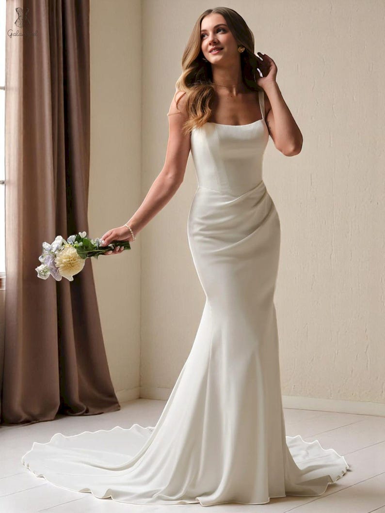 K&ouml;nnte beinhalten: Ein wei&szlig;es, bodenlanges Hochzeitskleid mit einer Meerjungfrau-Silhouette. Das Kleid hat einen quadratischen Ausschnitt, Spaghettitr&auml;ger und ein drapiertes Oberteil. Die Schleppe verl&auml;uft hinter dem Kleid. Die Person h&auml;lt einen Blumenstrau&szlig;.