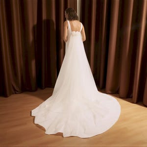 Puede incluir: Un vestido de novia blanco con una larga cola. El vestido tiene un dise&ntilde;o sencillo con una falda fluida y una espalda detallada. La novia est&aacute; de espaldas a la c&aacute;mara frente a cortinas marrones.