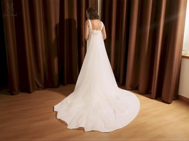 Puede incluir: Un vestido de novia blanco con una larga cola. El vestido tiene un dise&ntilde;o sencillo con una falda fluida y una espalda detallada. La novia est&aacute; de espaldas a la c&aacute;mara frente a cortinas marrones.