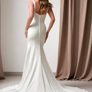 K&ouml;nnte beinhalten: Ein wei&szlig;es Brautkleid mit einer figurbetonten Silhouette und einer langen Schleppe. Das Kleid hat einen quadratischen Ausschnitt, Spaghettitr&auml;ger und eine Reihe von Kn&ouml;pfen am R&uuml;cken. Das Kleid ist aus einem glatten, flie&szlig;enden Stoff gefertigt.